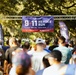 A large crowd of participants is gathered at the starting area of the '9/11 Heroes Run,' an annual commemorative race organized by the Travis Manion Foundation. The event is held outdoors in a park-like setting with large oak trees providing shade. CVS Health is identified as the national presenting sponsor on the main banner.