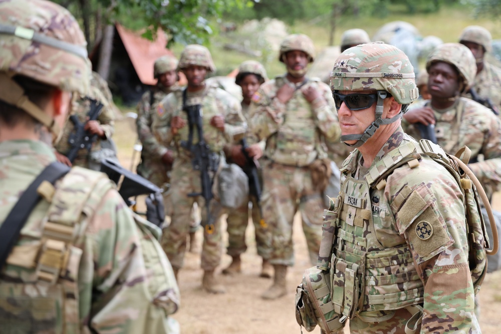 A group of military personnel in OCP (Operational Camouflage Pattern) uniforms are gathered in an outdoor field environment. A central figure, identifiable by a name tape reading 'PAZAK' and a two-star rank insignia on the helmet, appears to be a Major General interacting with or observing soldiers. The setting suggests a field training exercise or operational briefing.