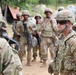 A group of military personnel in OCP (Operational Camouflage Pattern) uniforms are gathered in an outdoor field environment. A central figure, identifiable by a name tape reading 'PAZAK' and a two-star rank insignia on the helmet, appears to be a Major General interacting with or observing soldiers. The setting suggests a field training exercise or operational briefing.
