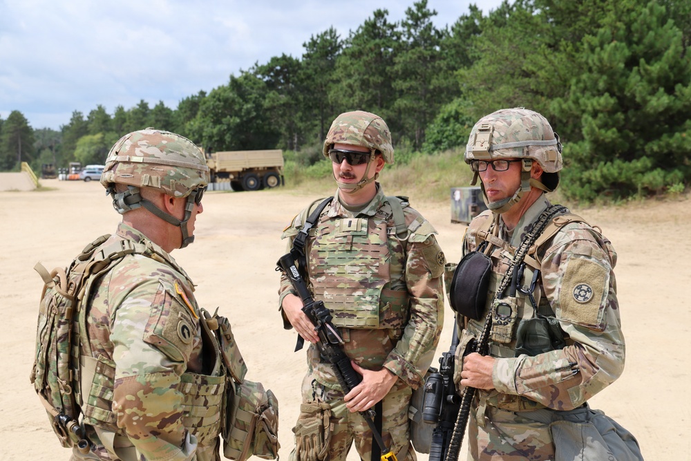 Three U.S. Army soldiers in full combat gear are engaged in a conversation at what appears to be a military training area or forward operating base. All three are wearing Operational Camouflage Pattern (OCP) uniforms, tactical vests, and helmets. The setting includes sandy terrain, military vehicles, and a tree line in the background.
