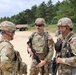 Three U.S. Army soldiers in full combat gear are engaged in a conversation at what appears to be a military training area or forward operating base. All three are wearing Operational Camouflage Pattern (OCP) uniforms, tactical vests, and helmets. The setting includes sandy terrain, military vehicles, and a tree line in the background.