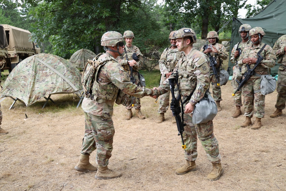 A group of U.S. Army soldiers in Operational Camouflage Pattern (OCP) uniforms are gathered in an outdoor field training environment. Two soldiers in the foreground are shaking hands, suggesting a recognition, greeting, or award ceremony. The setting includes military tents, a tactical vehicle, and wooded terrain consistent with a field exercise or training area.