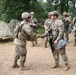 A group of U.S. Army soldiers in Operational Camouflage Pattern (OCP) uniforms are gathered in an outdoor field training environment. Two soldiers in the foreground are shaking hands, suggesting a recognition, greeting, or award ceremony. The setting includes military tents, a tactical vehicle, and wooded terrain consistent with a field exercise or training area.