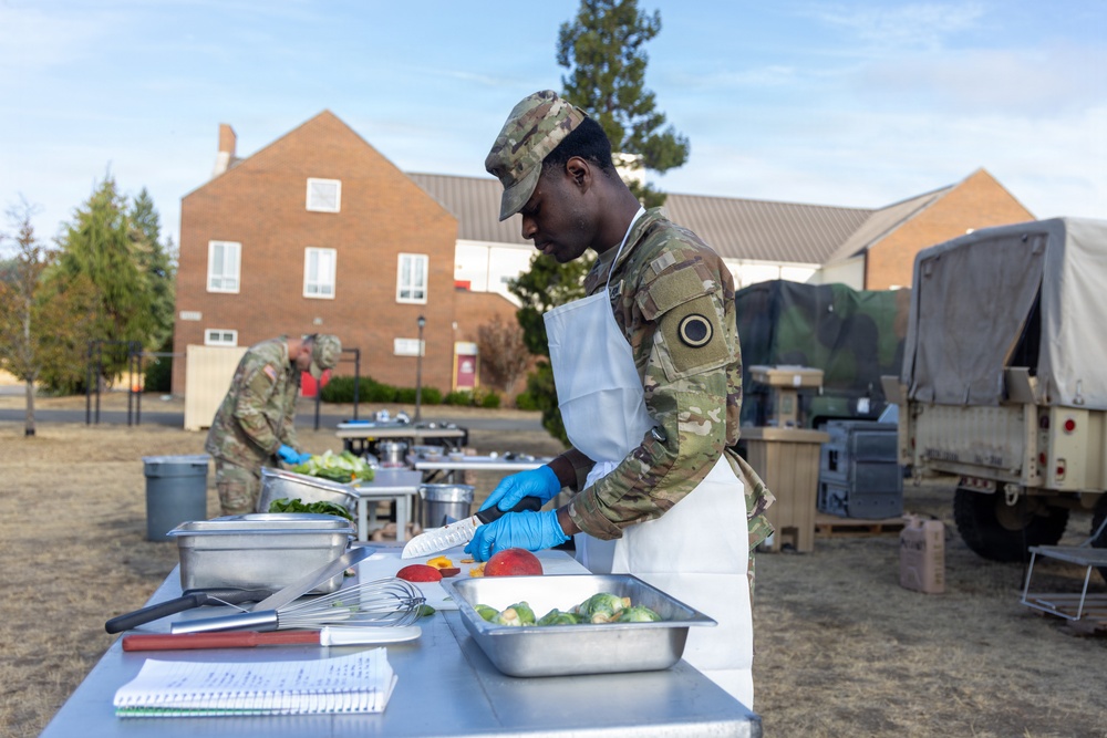 Two military personnel in camouflage uniforms and white aprons are preparing food outdoors on stainless steel tables, likely during a field cooking exercise or competition. The foreground soldier is cutting fruit while wearing blue nitrile gloves, with various cooking utensils and vegetables visible on the table. The setting appears to be a military installation with brick buildings and a military vehicle visible in the background.