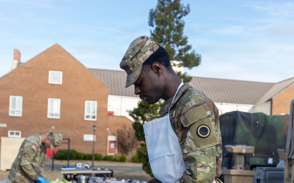 Two military personnel in camouflage uniforms and white aprons are preparing food outdoors on stainless steel tables, likely during a field cooking exercise or competition. The foreground soldier is cutting fruit while wearing blue nitrile gloves, with various cooking utensils and vegetables visible on the table. The setting appears to be a military installation with brick buildings and a military vehicle visible in the background.