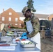 Two military personnel in camouflage uniforms and white aprons are preparing food outdoors on stainless steel tables, likely during a field cooking exercise or competition. The foreground soldier is cutting fruit while wearing blue nitrile gloves, with various cooking utensils and vegetables visible on the table. The setting appears to be a military installation with brick buildings and a military vehicle visible in the background.