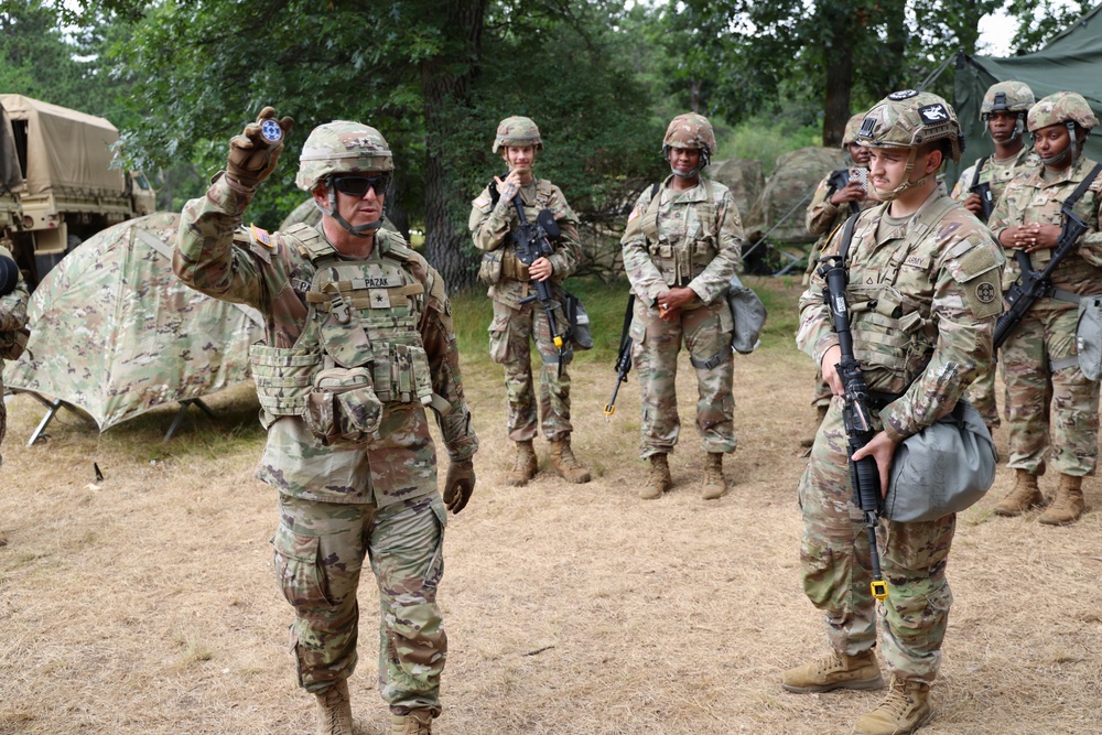 A group of U.S. Army soldiers in OCP (Operational Camouflage Pattern) uniforms are gathered in an outdoor field training environment. The soldier in the foreground, identified by name tape as 'PAZAK,' is holding up a small object — likely a coin or token — possibly presenting an award or making a point during a debrief. Several soldiers are carrying rifles and wearing full combat gear including helmets and plate carriers.
