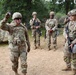 A group of U.S. Army soldiers in OCP (Operational Camouflage Pattern) uniforms are gathered in an outdoor field training environment. The soldier in the foreground, identified by name tape as 'PAZAK,' is holding up a small object — likely a coin or token — possibly presenting an award or making a point during a debrief. Several soldiers are carrying rifles and wearing full combat gear including helmets and plate carriers.