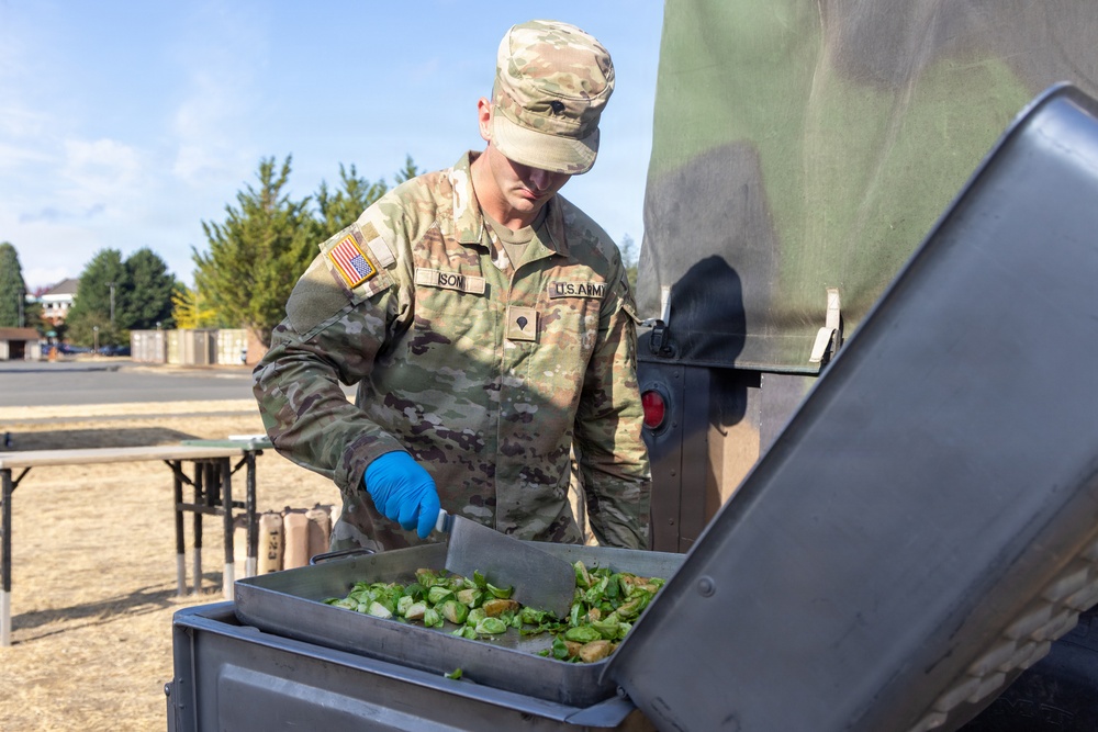 A U.S. Army soldier in OCP (Operational Camouflage Pattern) uniform is preparing food outdoors, handling what appears to be chopped green vegetables in a metal tray. The soldier is wearing blue nitrile gloves and using a spatula or scraper tool, suggesting field kitchen or outdoor cooking operations. The setting appears to be a military installation or training area based on the surroundings.