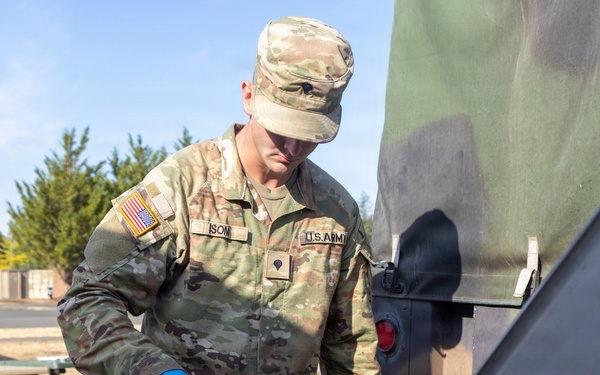 A U.S. Army soldier in OCP (Operational Camouflage Pattern) uniform is preparing food outdoors, handling what appears to be chopped green vegetables in a metal tray. The soldier is wearing blue nitrile gloves and using a spatula or scraper tool, suggesting field kitchen or outdoor cooking operations. The setting appears to be a military installation or training area based on the surroundings.