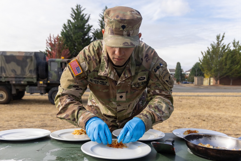 A U.S. Army soldier named Reyes Cruz is carefully plating food onto white dishes outdoors on a military installation. The soldier is wearing full OCP (Operational Camouflage Pattern) uniform with blue latex gloves, suggesting participation in a military culinary competition or field food service event. Multiple white plates are arranged on a green table, with what appears to be a shredded or fried food item being portioned out.