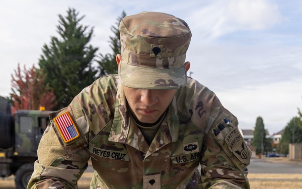 A U.S. Army soldier named Reyes Cruz is carefully plating food onto white dishes outdoors on a military installation. The soldier is wearing full OCP (Operational Camouflage Pattern) uniform with blue latex gloves, suggesting participation in a military culinary competition or field food service event. Multiple white plates are arranged on a green table, with what appears to be a shredded or fried food item being portioned out.