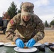 A U.S. Army soldier named Reyes Cruz is carefully plating food onto white dishes outdoors on a military installation. The soldier is wearing full OCP (Operational Camouflage Pattern) uniform with blue latex gloves, suggesting participation in a military culinary competition or field food service event. Multiple white plates are arranged on a green table, with what appears to be a shredded or fried food item being portioned out.
