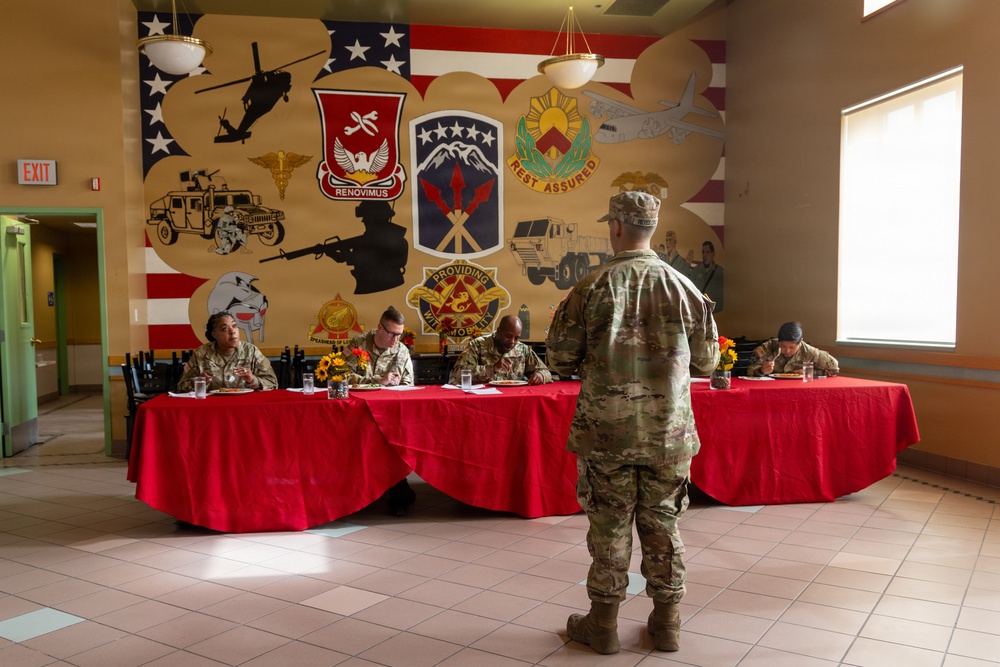 A military dining facility or mess hall setting where uniformed soldiers are seated at a long table covered with a red tablecloth, appearing to be engaged in a formal meal or evaluation event. A soldier in camouflage uniform stands with their back to the camera, facing the seated panel. The background features a large patriotic mural displaying multiple U.S. Army unit insignia, including patches reading 'Renovimus' and 'Rest Assured,' along with military vehicle and aircraft silhouettes over an American flag backdrop.
