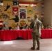 A military dining facility or mess hall setting where uniformed soldiers are seated at a long table covered with a red tablecloth, appearing to be engaged in a formal meal or evaluation event. A soldier in camouflage uniform stands with their back to the camera, facing the seated panel. The background features a large patriotic mural displaying multiple U.S. Army unit insignia, including patches reading 'Renovimus' and 'Rest Assured,' along with military vehicle and aircraft silhouettes over an American flag backdrop.