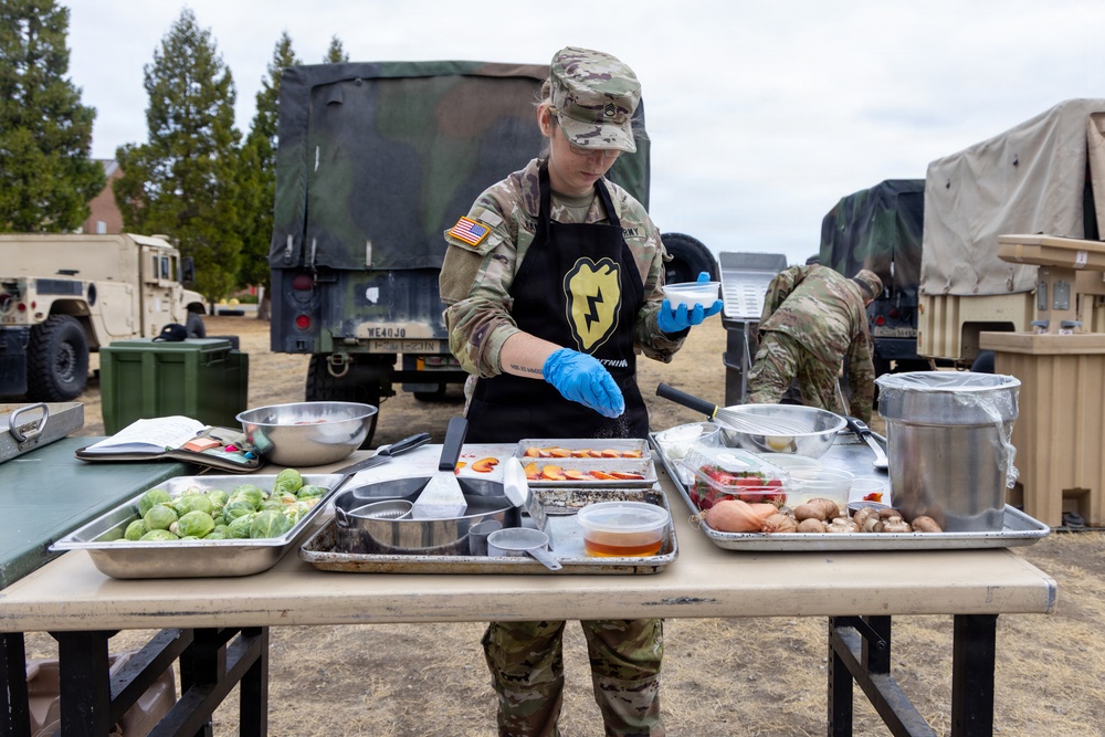 A U.S. Army soldier in camouflage uniform and black apron prepares food at an outdoor field kitchen setup. The soldier is wearing blue food-handling gloves and seasoning ingredients arranged on metal sheet pans on a folding table. Military vehicles and equipment are visible in the background, suggesting a field training or operational environment.