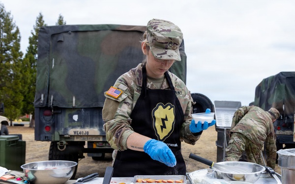 A U.S. Army soldier in camouflage uniform and black apron prepares food at an outdoor field kitchen setup. The soldier is wearing blue food-handling gloves and seasoning ingredients arranged on metal sheet pans on a folding table. Military vehicles and equipment are visible in the background, suggesting a field training or operational environment.