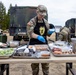 A U.S. Army soldier in camouflage uniform and black apron prepares food at an outdoor field kitchen setup. The soldier is wearing blue food-handling gloves and seasoning ingredients arranged on metal sheet pans on a folding table. Military vehicles and equipment are visible in the background, suggesting a field training or operational environment.