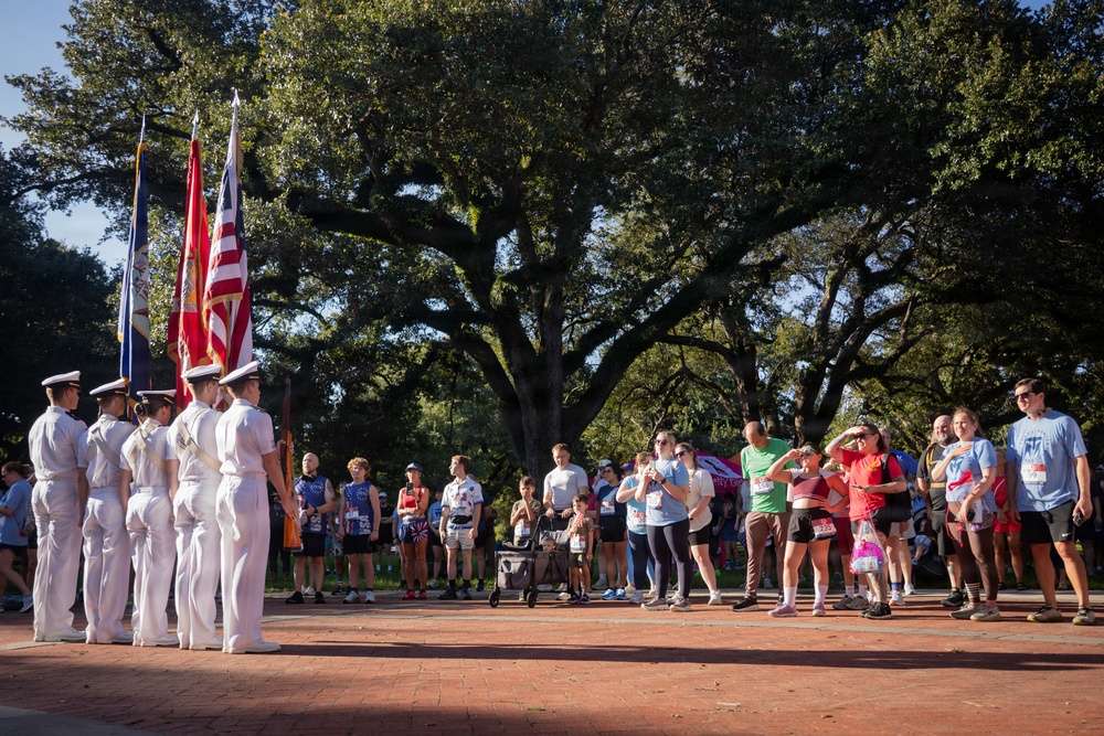 A color guard ceremony is taking place outdoors on a brick plaza, with uniformed personnel holding multiple flags including the American flag. A crowd of civilians, many wearing race bibs, stands watching the ceremony, suggesting this is the opening of a running race or similar public event. The setting features large oak trees providing a canopy backdrop in what appears to be a park or public square.
