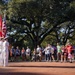 A color guard ceremony is taking place outdoors on a brick plaza, with uniformed personnel holding multiple flags including the American flag. A crowd of civilians, many wearing race bibs, stands watching the ceremony, suggesting this is the opening of a running race or similar public event. The setting features large oak trees providing a canopy backdrop in what appears to be a park or public square.