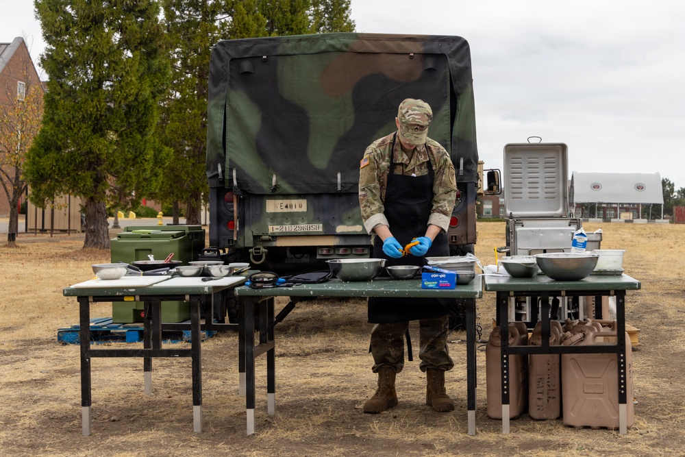 A soldier in camouflage uniform and black apron is preparing food at an outdoor field kitchen setup. They are working at folding tables covered with stainless steel bowls, pans, and cooking equipment, positioned in front of a military tactical vehicle. The setting appears to be a military base or training area, likely a field cooking exercise or competition.