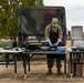 A soldier in camouflage uniform and black apron is preparing food at an outdoor field kitchen setup. They are working at folding tables covered with stainless steel bowls, pans, and cooking equipment, positioned in front of a military tactical vehicle. The setting appears to be a military base or training area, likely a field cooking exercise or competition.