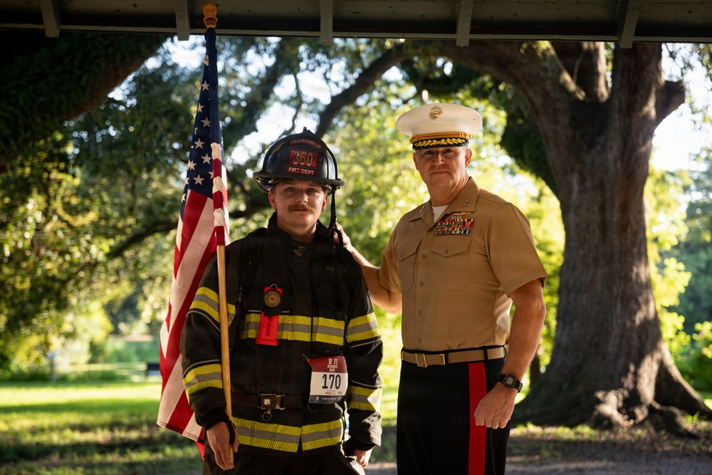 Two individuals pose together outdoors beneath a covered structure with large oak trees in the background. One person is dressed in full firefighter turnout gear, wearing a black helmet marked '360 Fire Dept' and holding an American flag, with a race bib numbered '170' suggesting a 9/11 memorial run. The other individual is wearing a United States Marine Corps dress uniform with multiple service ribbons and the distinctive white cover (hat).