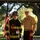 Audubon Park Hosts the 2026 New Orleans 9/11 Heroes Run Two individuals pose together outdoors beneath a covered structure with large oak trees in the background. One person is dressed in full firefighter turnout gear, wearing a black helmet marked '360 Fire Dept' and holding an American flag, with a race bib numbered '170' suggesting a 9/11 memorial run. The other individual is wearing a United States Marine Corps dress uniform with multiple service ribbons and the distinctive white cover (hat).