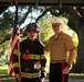 Two individuals pose together outdoors beneath a covered structure with large oak trees in the background. One person is dressed in full firefighter turnout gear, wearing a black helmet marked '360 Fire Dept' and holding an American flag, with a race bib numbered '170' suggesting a 9/11 memorial run. The other individual is wearing a United States Marine Corps dress uniform with multiple service ribbons and the distinctive white cover (hat).