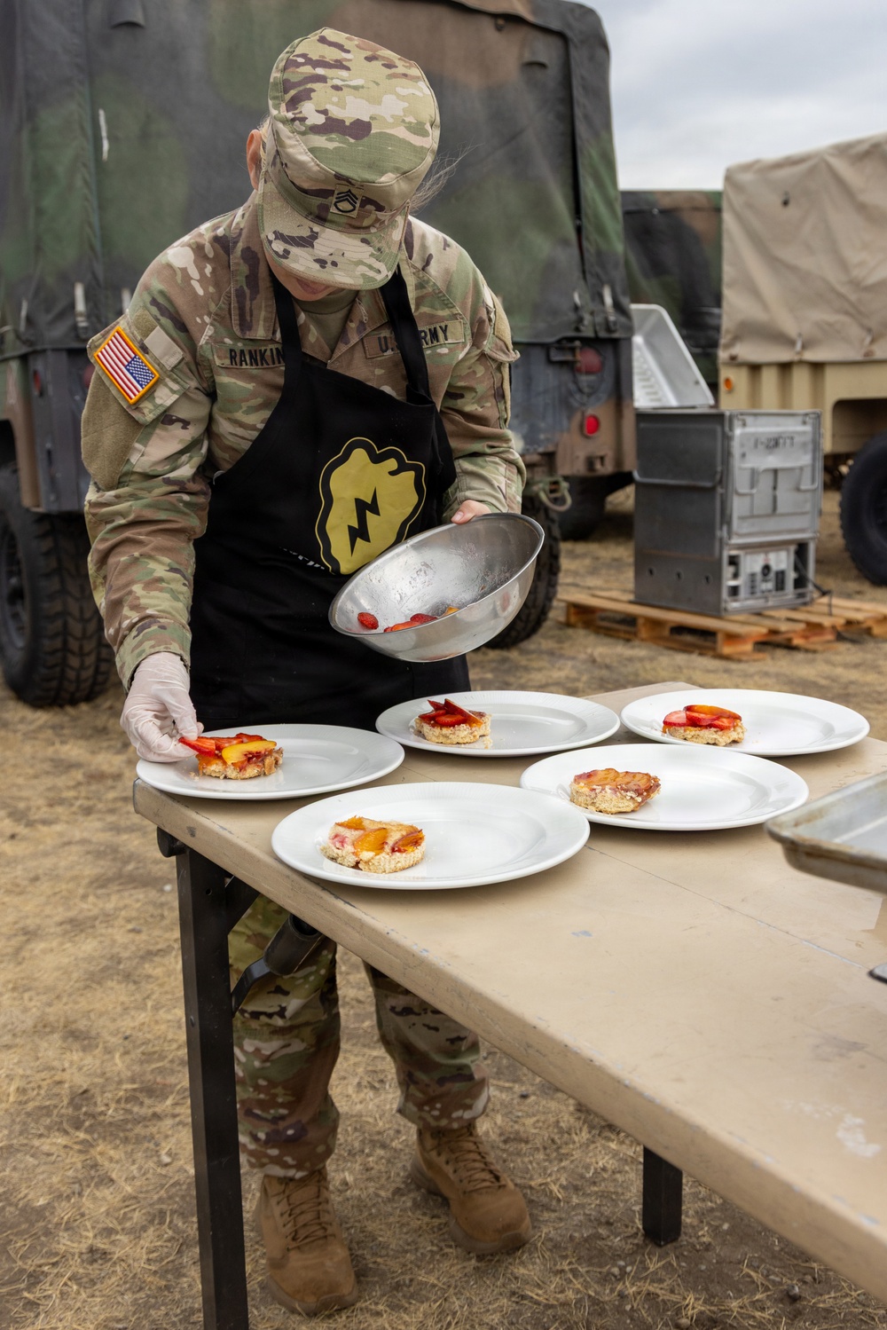 A U.S. Army soldier named Rankin, wearing OCP camouflage uniform with a black apron bearing a unit insignia (lightning bolt on an acorn/shield patch), is plating food items onto white plates at an outdoor field setting. The soldier appears to be participating in a military culinary competition or field cooking exercise, carefully arranging fruit-topped pastry or cake portions on five white plates. Military vehicles and equipment are visible in the background.