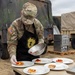 A U.S. Army soldier named Rankin, wearing OCP camouflage uniform with a black apron bearing a unit insignia (lightning bolt on an acorn/shield patch), is plating food items onto white plates at an outdoor field setting. The soldier appears to be participating in a military culinary competition or field cooking exercise, carefully arranging fruit-topped pastry or cake portions on five white plates. Military vehicles and equipment are visible in the background.