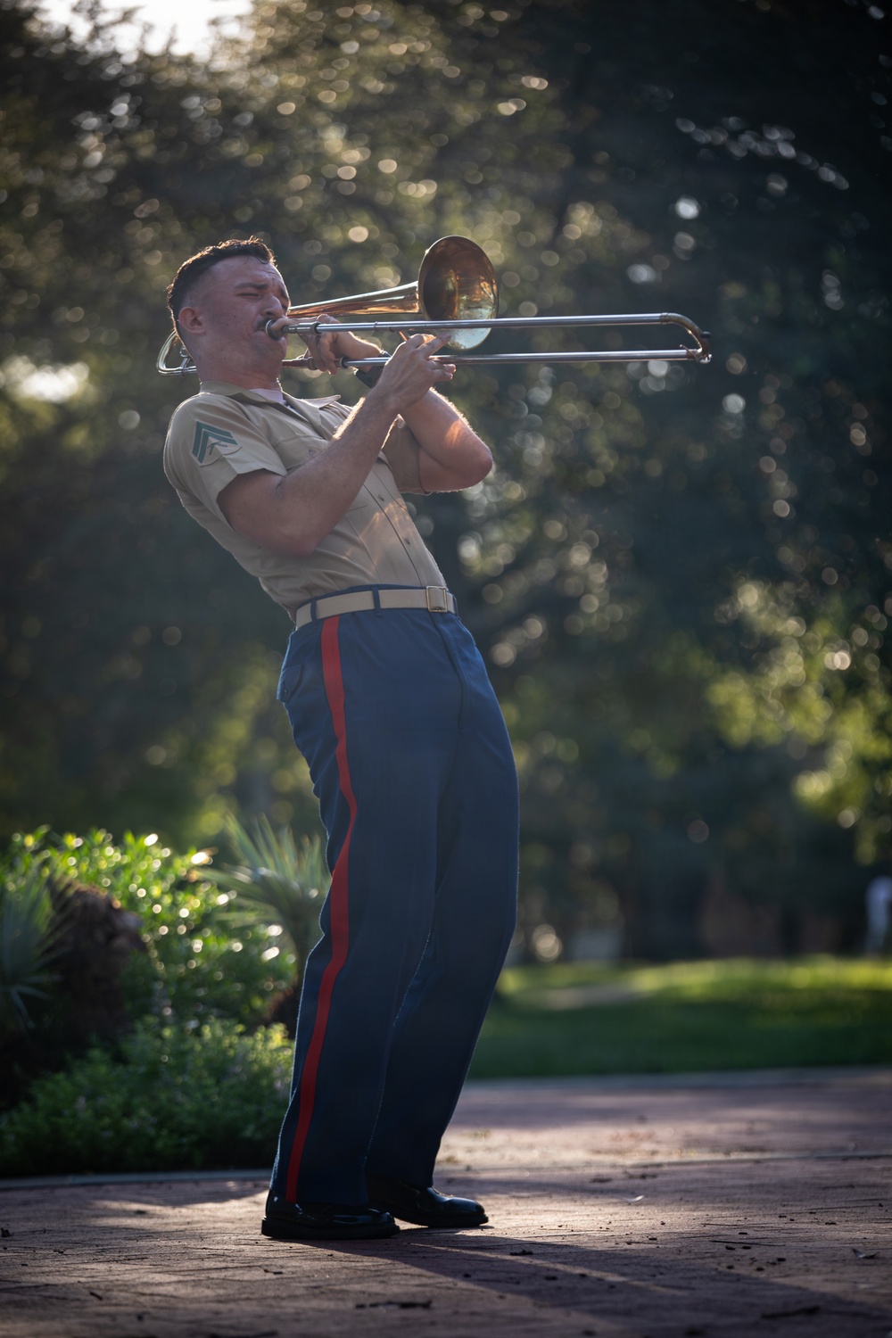 A U.S. Marine in service uniform is playing a trombone outdoors on a brick pathway, leaning back with apparent intensity while performing. He is wearing the characteristic khaki shirt and blue trousers with a red 'blood stripe' down the leg, consistent with USMC dress/service attire. The background features lush, bokeh-blurred trees illuminated by warm, directional sunlight.