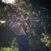 A U.S. Marine in service uniform is playing a trombone outdoors on a brick pathway, leaning back with apparent intensity while performing. He is wearing the characteristic khaki shirt and blue trousers with a red 'blood stripe' down the leg, consistent with USMC dress/service attire. The background features lush, bokeh-blurred trees illuminated by warm, directional sunlight.