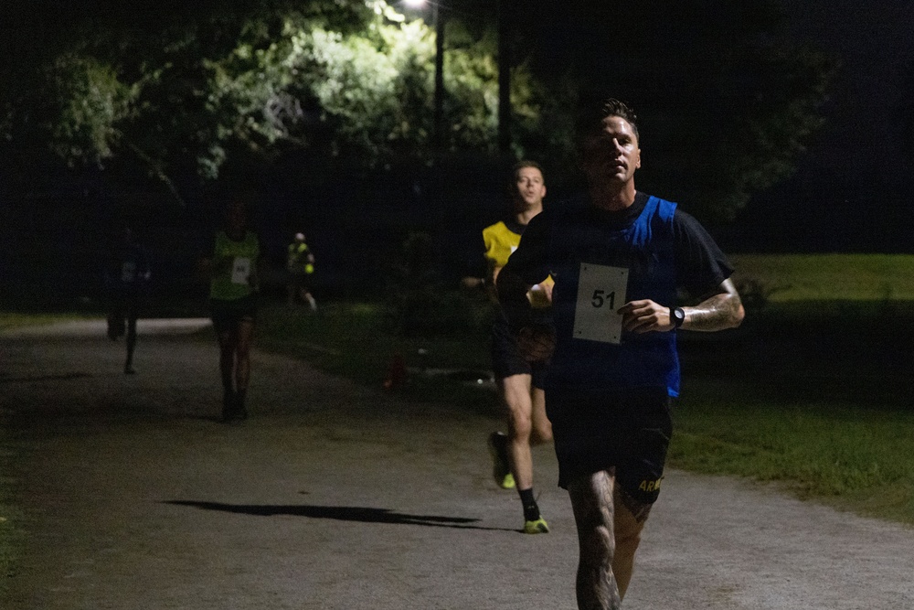 A nighttime running race captured on a paved path in a park-like setting. The lead runner, wearing bib number 51, a blue vest over a black shirt, and black shorts with 'ARM' visible, is in sharp focus in the foreground. Several other runners are visible behind him, illuminated by overhead street lighting.