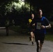 A nighttime running race captured on a paved path in a park-like setting. The lead runner, wearing bib number 51, a blue vest over a black shirt, and black shorts with 'ARM' visible, is in sharp focus in the foreground. Several other runners are visible behind him, illuminated by overhead street lighting.