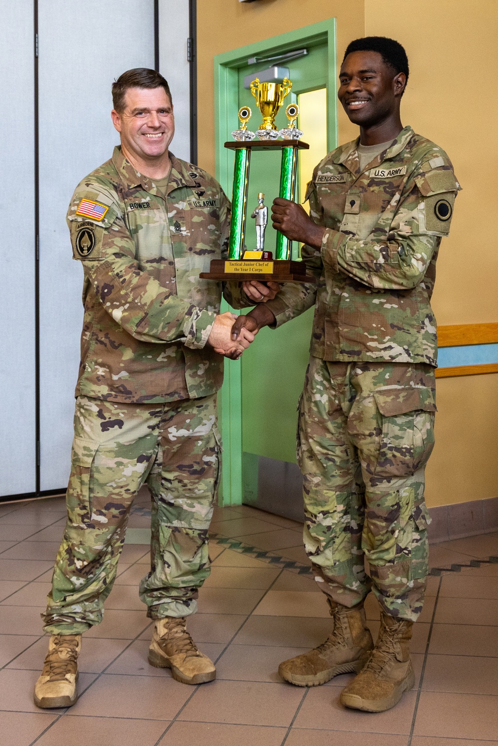 Two U.S. Army soldiers in OCP (Operational Camouflage Pattern) uniforms are shaking hands while jointly holding a large trophy. The soldier on the right, identified by his name tape as 'Henderson,' appears to be receiving the award from the soldier on the left, identified as 'Bower.' The trophy's plaque reads 'Tactical Junior Chef of the Year I Corps.'