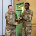 Two U.S. Army soldiers in OCP (Operational Camouflage Pattern) uniforms are shaking hands while jointly holding a large trophy. The soldier on the right, identified by his name tape as 'Henderson,' appears to be receiving the award from the soldier on the left, identified as 'Bower.' The trophy's plaque reads 'Tactical Junior Chef of the Year I Corps.'