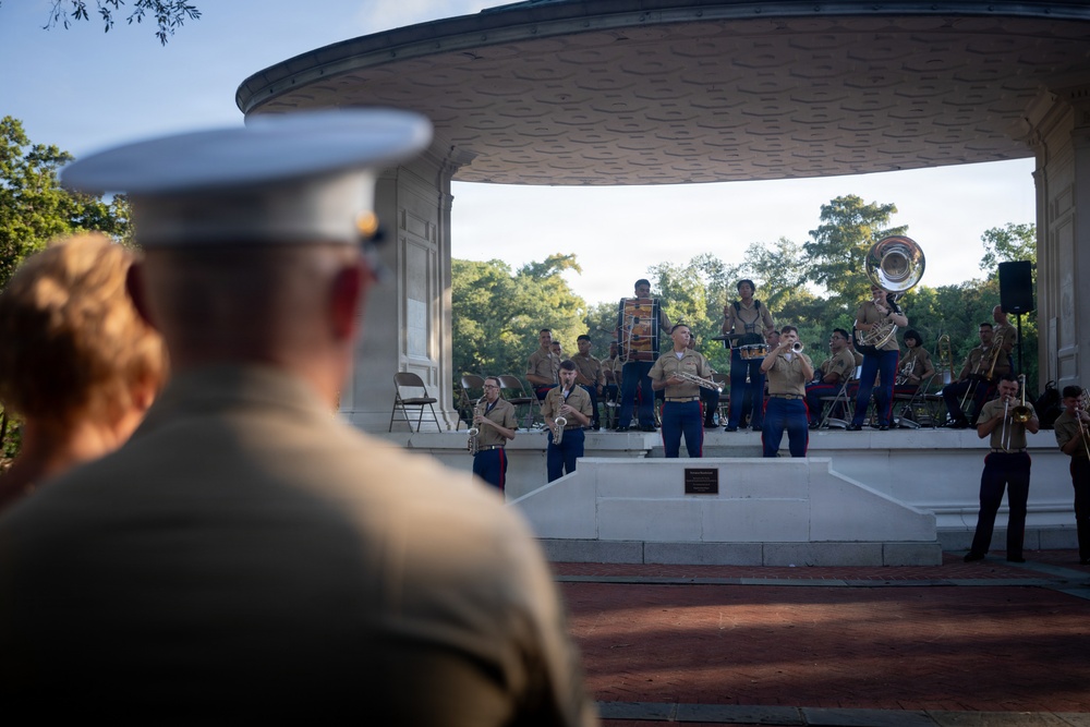 A U.S. Marine Corps band performs at an outdoor bandstand/pavilion, observed by a Marine officer in the foreground wearing a white dress cover. The musicians are dressed in tan/khaki utility uniforms and are playing a variety of brass and percussion instruments. The setting appears to be a public park or ceremonial outdoor venue during evening hours.