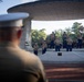 A U.S. Marine Corps band performs at an outdoor bandstand/pavilion, observed by a Marine officer in the foreground wearing a white dress cover. The musicians are dressed in tan/khaki utility uniforms and are playing a variety of brass and percussion instruments. The setting appears to be a public park or ceremonial outdoor venue during evening hours.