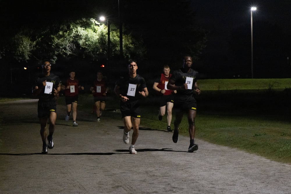 A group of military personnel, identifiable by 'ARMY' branded athletic wear, are participating in a nighttime running event or physical fitness test on a paved path. Runners wear numbered bibs (visible numbers include 27, 28, and 30), suggesting a timed or competitive assessment. The scene is illuminated by overhead street lights against a dark sky.