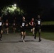 A group of military personnel, identifiable by 'ARMY' branded athletic wear, are participating in a nighttime running event or physical fitness test on a paved path. Runners wear numbered bibs (visible numbers include 27, 28, and 30), suggesting a timed or competitive assessment. The scene is illuminated by overhead street lights against a dark sky.