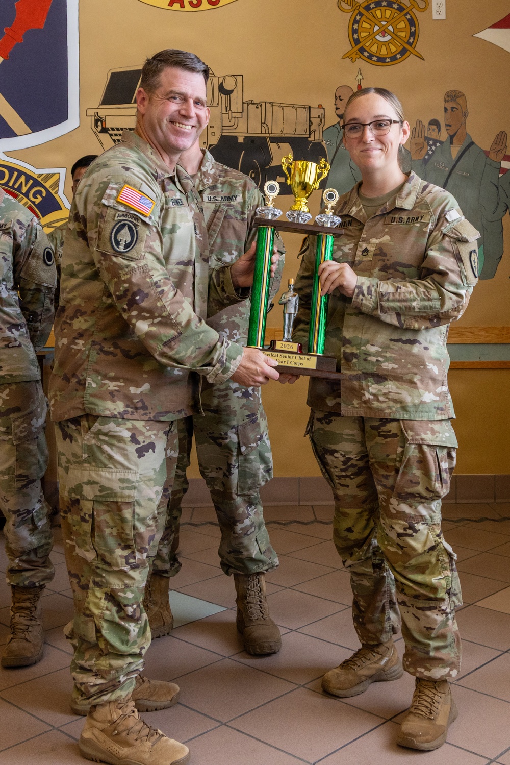 Two U.S. Army soldiers in OCP (Operational Camouflage Pattern) uniforms are jointly holding a trophy during what appears to be an award ceremony. The trophy plaque reads '2026 Tactical Senior Chef of the Year I Corps,' indicating a culinary excellence recognition. The ceremony is taking place indoors in front of a painted mural featuring military insignia and imagery.