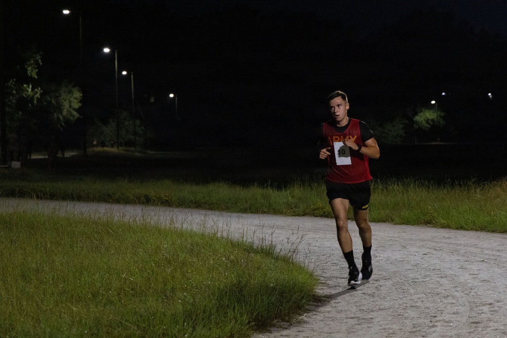 A male runner wearing a red 'ARMY' vest with race bib number 10 is running along a paved path at night. The scene is illuminated by street lights in what appears to be a military base or park setting. The runner is mid-stride, wearing black shorts, black socks, and running shoes.