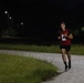 A male runner wearing a red 'ARMY' vest with race bib number 10 is running along a paved path at night. The scene is illuminated by street lights in what appears to be a military base or park setting. The runner is mid-stride, wearing black shorts, black socks, and running shoes.