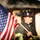 Audubon Park Hosts the 2026 New Orleans 9/11 Heroes Run A firefighter from Prairieville Fire Department, wearing a black helmet numbered 360, stands holding an American flag on a wooden pole. The subject is dressed in full turnout gear with yellow reflective striping and appears to be at an outdoor location with a blurred background. The image has a solemn, patriotic tone suggesting a ceremonial or commemorative event.