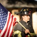 A firefighter from Prairieville Fire Department, wearing a black helmet numbered 360, stands holding an American flag on a wooden pole. The subject is dressed in full turnout gear with yellow reflective striping and appears to be at an outdoor location with a blurred background. The image has a solemn, patriotic tone suggesting a ceremonial or commemorative event.