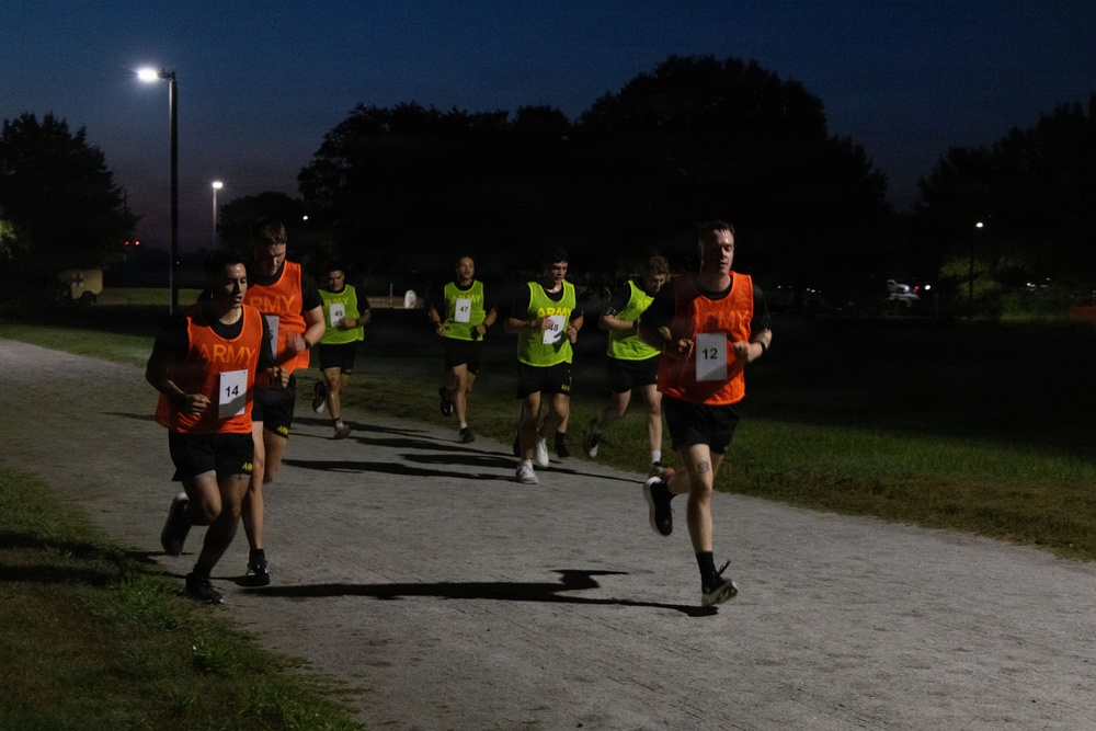 A group of runners wearing 'ARMY' branded bibs in orange and neon yellow/green are participating in a nighttime running event on a paved path. The scene is illuminated by overhead street lighting, with trees and parked vehicles visible in the background. Visible bib numbers include 12, 14, 47, 48, and 49, suggesting a structured competitive or fitness assessment event.