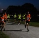 A group of runners wearing 'ARMY' branded bibs in orange and neon yellow/green are participating in a nighttime running event on a paved path. The scene is illuminated by overhead street lighting, with trees and parked vehicles visible in the background. Visible bib numbers include 12, 14, 47, 48, and 49, suggesting a structured competitive or fitness assessment event.