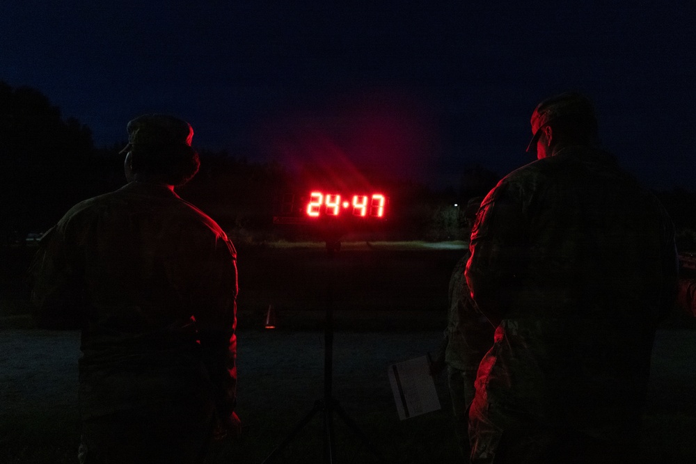 Two individuals in military camouflage uniforms are standing in darkness, facing a red LED digital display showing the time or countdown of 24:47. The scene appears to be an outdoor nighttime military training or fitness event, likely a physical fitness test or timed event. The red digital clock on a stand is the primary light source illuminating the silhouetted figures.