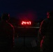 Two individuals in military camouflage uniforms are standing in darkness, facing a red LED digital display showing the time or countdown of 24:47. The scene appears to be an outdoor nighttime military training or fitness event, likely a physical fitness test or timed event. The red digital clock on a stand is the primary light source illuminating the silhouetted figures.